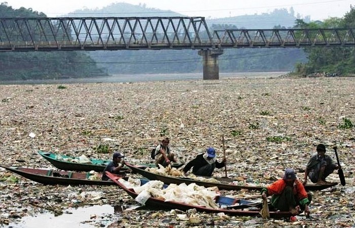 Мужчины на лодке ищут пластиковые бутылки среди груд мусора в реке.